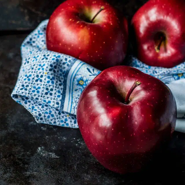 Three red apples sitting in a bowl with a blue floral towel