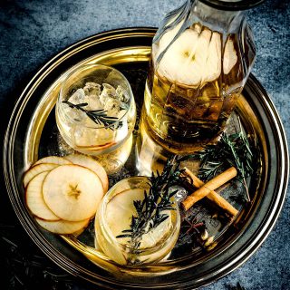 Mulled cider served in a glass carafe with glasses, cinnamon sticks, rosemary sprigs, and sliced apples