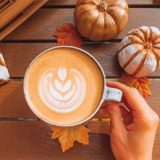 Hand holding a mug of pumpkin spice latte with latte art on a wooden table next to small pumpkins