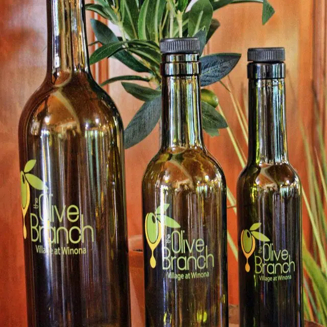 Three extra virgin olive oil bottles with The Olive Branch logo on them in varying sizes on top of a wooden barrel