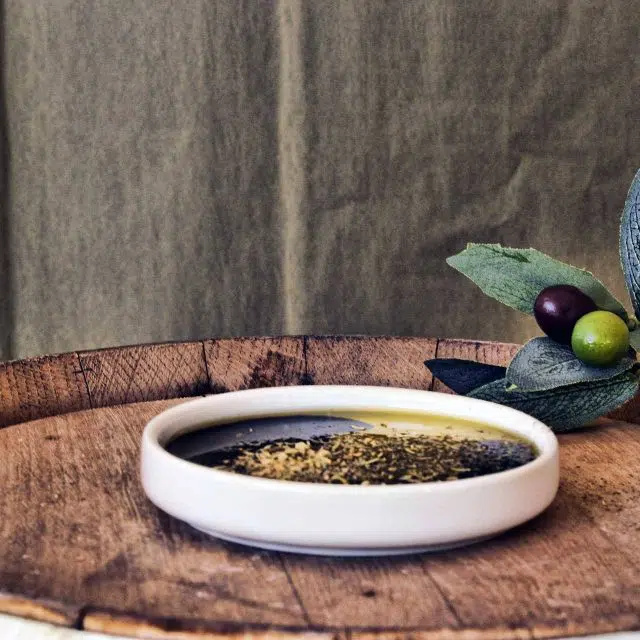 Shallow white bowl of olive oil, vinegar, and herbs sitting on the top of a wooden barrel