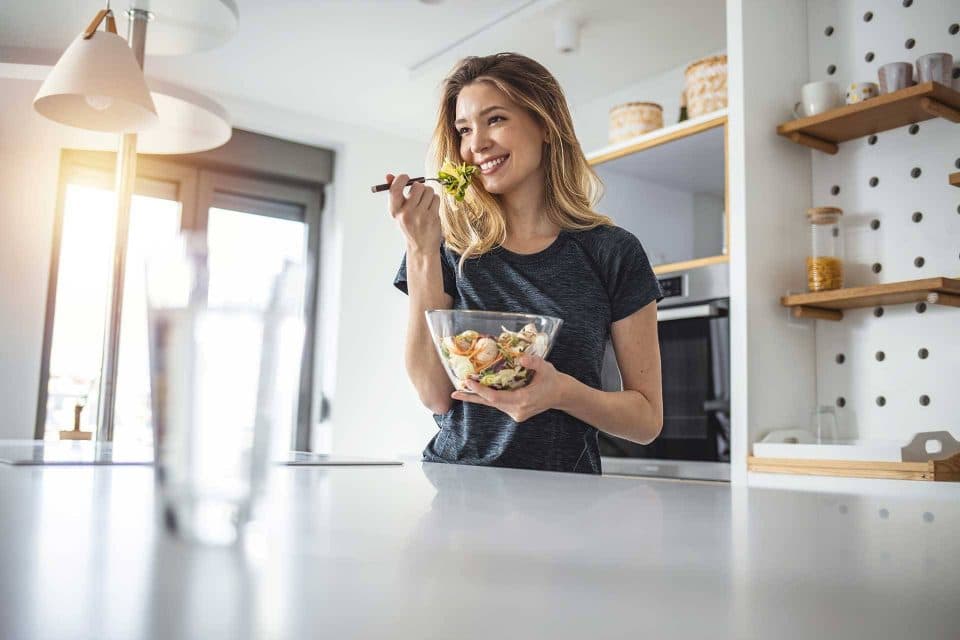 White woman smiling as she eats salad out of a glass bowl in a kitchen