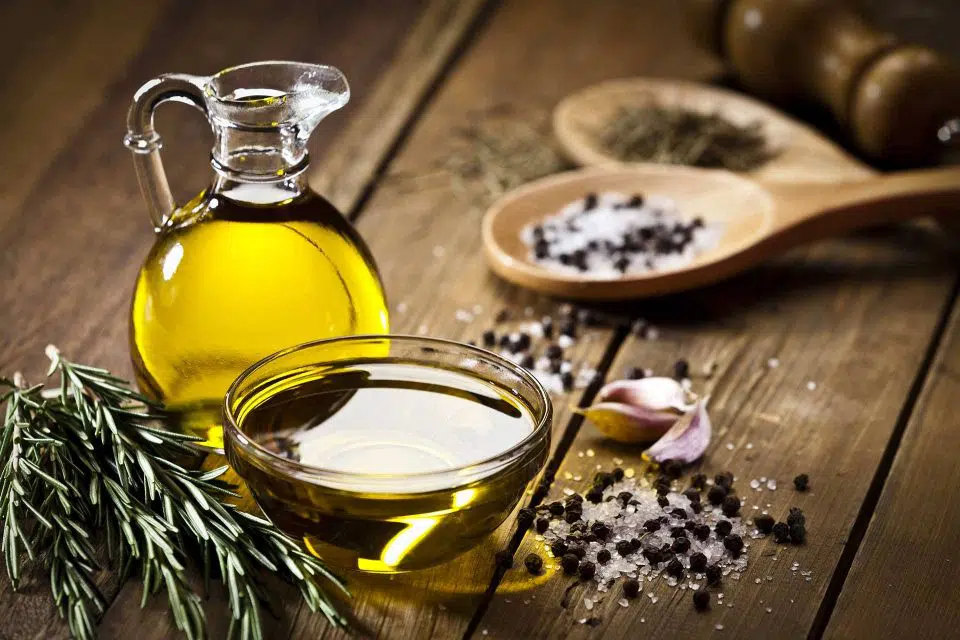 Glass carafe full of extra virgin olive oil next to a glass bowl, fresh rosemary sprigs, and wooden spoons full of salt and pepper