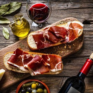Slices of bread topped with proscuitto served with olives, pickles, parmesan cheese, extra virgin olive oil and red wine on a wooden board