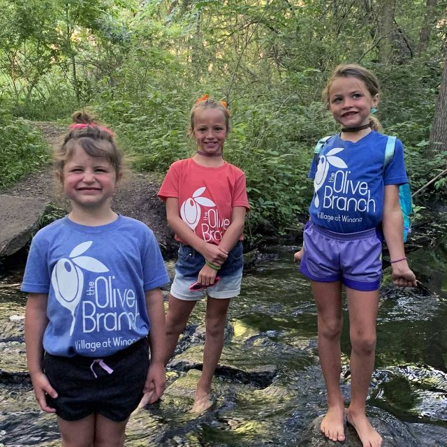 Three children in Olive Branch t-shirts standing in a creek smiling at the camera